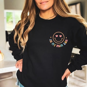 Puede incluir: Sudadera negra con un gr&aacute;fico de arco&iacute;ris que dice "In My Mom Era" y una carita sonriente con corazones por ojos.