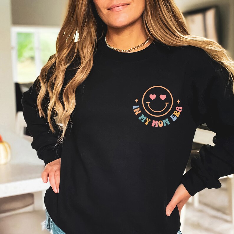 Puede incluir: Sudadera negra con un gr&aacute;fico de arco&iacute;ris que dice "In My Mom Era" y una carita sonriente con corazones por ojos.