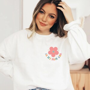 Puede incluir: Sudadera blanca con un gr&aacute;fico de flor rosa y el texto "In My Mom Era" en colores arco&iacute;ris.