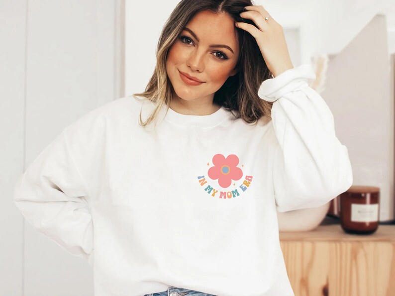 Puede incluir: Sudadera blanca con un gr&aacute;fico de flor rosa y el texto "In My Mom Era" en colores arco&iacute;ris.