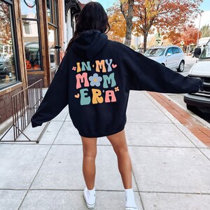 Puede incluir: Sudadera con capucha negra con el texto "In My Mom Era" en letras retro de colores con un gr&aacute;fico de flores.