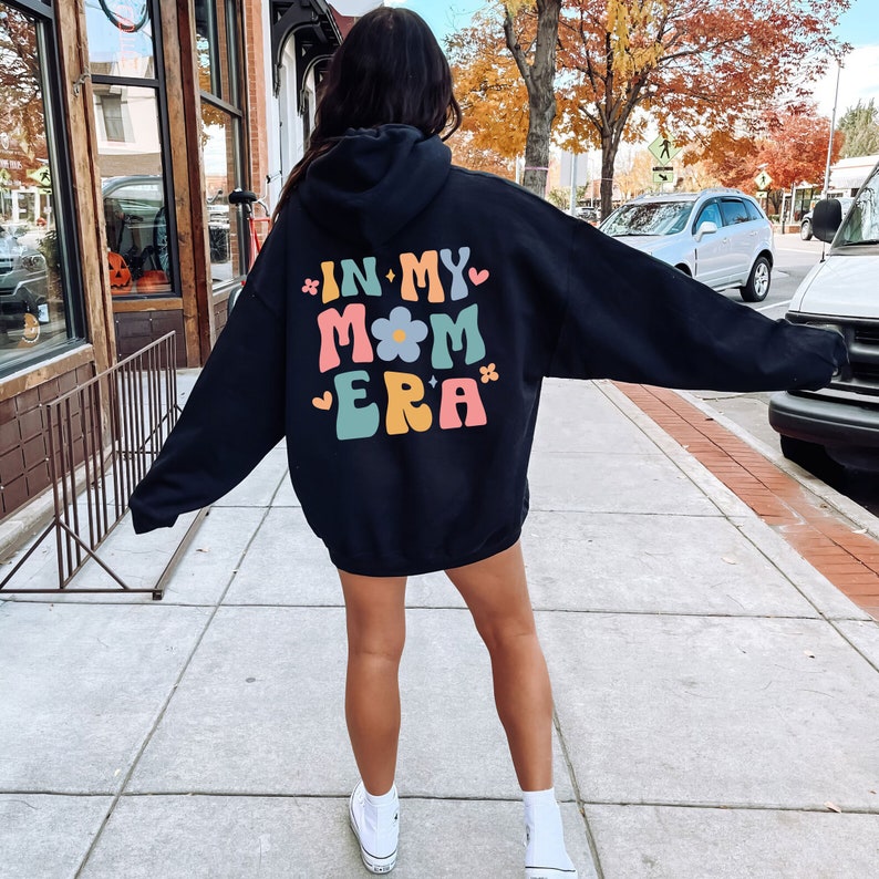 Puede incluir: Sudadera con capucha negra con el texto "In My Mom Era" en letras retro de colores con un gr&aacute;fico de flores.