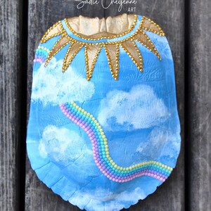 Könnte beinhalten: Eine blaue Schildkrötenschale, die mit einer Himmelsszene, weißen Wolken und einem Regenbogen aus bunten Perlen bemalt ist. Die Oberseite der Schale hat ein goldenes Sonnenstrahlen-Design mit goldenen Perlen.