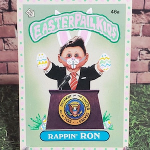 May include: A green and pink trading card with a cartoon image of a man wearing a bunny suit and holding Easter eggs. The card is titled "Easterpal Kids" and features the text "Rappin' Ron" at the bottom. The card number is 46a.