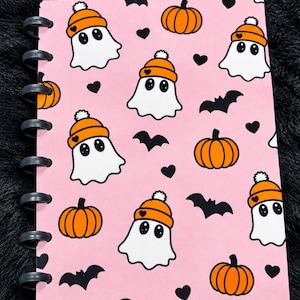 May include: A disc-bound notebook with a light pink cover featuring a repeating pattern of white ghosts wearing orange beanies, orange pumpkins, black bats, and small black hearts. The notebook has black plastic binding discs along the left side.