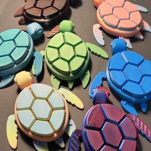 May include: A set of 7 colorful 3D printed turtle figurines. The turtles are all different colors, including brown, pink, green, blue, and teal. Each turtle has a detailed shell with a hexagonal pattern.
