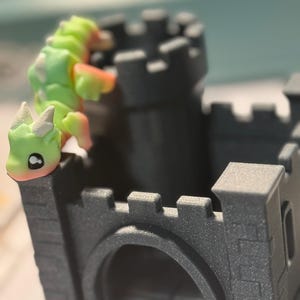 Puede incluir: Una figurita de dragón verde y naranja trepando por un juguete de castillo gris. El castillo tiene una pequeña puerta arqueada y una torre en la parte superior.