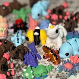 May include: A collection of colorful, small figurines, including a white unicorn, blue dolphin-like creatures, and brown bear-like figures with pink accents. Other figures include a yellow and black design, a green dinosaur, and a purple fish-like creature.