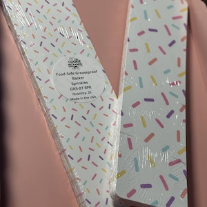 Puede incluir: Dos pilas de hojas de respaldo rectangulares a prueba de grasa y seguras para alimentos. Las hojas son blancas con un patrón de chispas de colores. El texto en el empaque dice "Food-Safe Greaseproof Backer Sprinkles GRS-27-SPR Quantity: 25 Made in the USA."