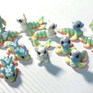 May include: A collection of small, colorful fantasy creature figurines. The figurines include various designs, such as dragons, caterpillar-like creatures, and small humanoid figures, in shades of blue, green, white, and peach.