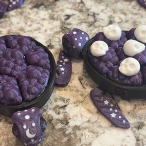 May include: Close-up of several purple and black decorative objects. One object is a small bowl filled with purple, textured material and topped with white skulls. Other objects include dice and star-shaped pieces.