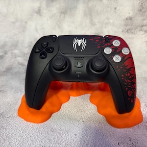 Puede incluir: Mando de PlayStation 5 negro y rojo con un logotipo de araña blanco y un soporte naranja. El mando tiene un cuerpo negro con detalles rojos y símbolos blancos. El soporte es de color naranja brillante.