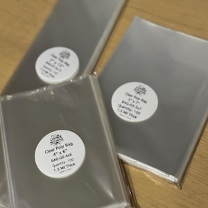 Pode incluir: Vários sacos de polietileno transparentes são exibidos, com etiquetas circulares brancas. As etiquetas dizem "Clear Poly Bag" com detalhes de tamanho e quantidade. Um saco é rotulado 10 x 15 cm, outro 13 x 18 cm. Os sacos têm 1,5 Mil de espessura.