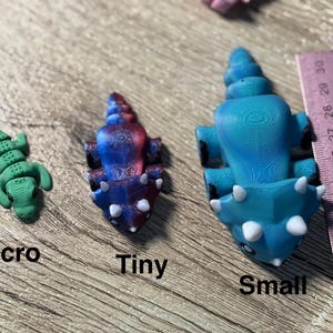 Pode incluir: Uma coleção de figuras de dinossauros impressas em 3D em vários tamanhos: micro, pequeno e pequeno. O dinossauro pequeno tem aproximadamente 6,35 cm de comprimento. As figuras são azuis, verdes, rosa e vermelhas, com detalhes brancos. As palavras "Micro", "Tiny" e "Small" são impressas abaixo.