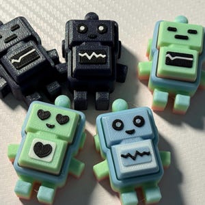 May include: A collection of colorful robot figurines in various shades of blue and green. Each robot has a unique face and design, with details like hearts and zig-zag patterns. The robots are small and appear to be 3D-printed.