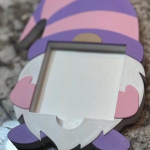 May include: A whimsical gnome-shaped picture frame. The frame features a striped purple and pink hat, white beard, and pink hands. The frame is designed to hold a small photo or artwork. The overall design is a playful and decorative item.