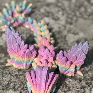 Könnte beinhalten: Eine farbenfrohe, mehrschichtige, schillernde Drachenfigur mit einem Regenbogen-Farbverlauf in Rosa, Gelb und Blau. Der Drache hat einen langen, segmentierten Schwanz und gefiederte Flügel.