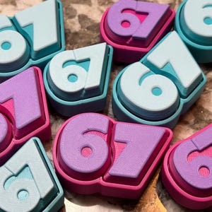 Op de afbeelding: Een verzameling kleurrijke 3D-geprinte cijfers 67. De cijfers hebben een gelaagd ontwerp met een lichtblauwe bovenlaag, een witte middenlaag en een turquoise of roze basis. De cijfers zijn verspreid over een oppervlak.