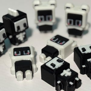Könnte beinhalten: Eine Sammlung kleiner, blockartiger Figuren in Schwarz und Weiß. Die Figuren haben ein Skelettdesign mit quadratischen Köpfen, rechteckigen Körpern und kleinen Füßen. Einige Figuren stehen aufrecht, andere liegen.