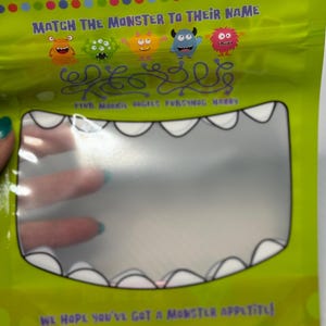 May include: A green plastic bag with a clear window shaped like a monster's mouth. The bag features colorful cartoon monsters and text that reads, "Munch these treats before the 'til monsters get 'em!" and "Match the monster to their name."