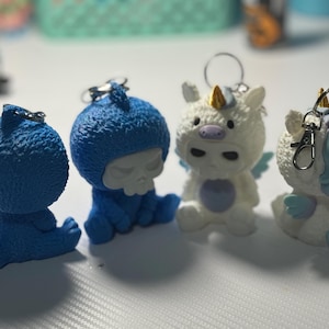 May include: Four whimsical keychains: a blue shark, a blue figure with a skull face, and two white unicorns with wings. The unicorn keychains have gold horns and blue accents. Each keychain has a silver ring for attaching to keys or bags.