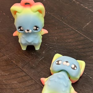 Op de afbeelding: Twee kleurrijke, 3D-geprinte beeldjes van een cartoon dier met grote ogen en kleine armen. De beeldjes zijn voornamelijk blauw, met accenten van geel, roze en groen. Eén beeldje staat rechtop, de andere ligt op zijn zij.