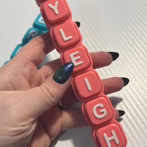 Op de afbeelding: Een roze naamlabel met de naam "Ryleigh" in witte letters. Het naamlabel is gemaakt van verbonden vierkante blokken. Een tweede blauw naamlabel is gedeeltelijk zichtbaar op de achtergrond.
