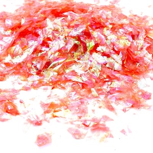 May include: A pile of iridescent pink and gold glitter flakes. The glitter is scattered on a white surface.