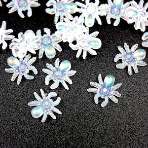 Puede incluir: Una colección de adornos iridiscentes con forma de araña. Estas piezas decorativas presentan un cuerpo transparente y brillante con toques de azul, morado y verde. Los abalorios de araña están dispersos sobre una superficie oscura y texturizada.