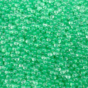 Puede incluir: Una imagen de primer plano de una gran cantidad de pequeñas cuentas verdes translúcidas. Las cuentas son redondas y tienen un ligero brillo iridiscente.