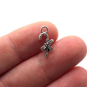 May include: A small, silver-coloured charm shaped like a candy cane with a bow. The charm is held between fingers against a white background. Suitable for jewellery making and crafting.