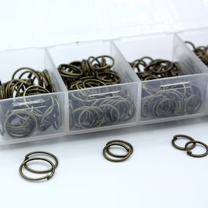 Könnte beinhalten: Eine transparente Plastik-Aufbewahrungsbox gefüllt mit kleinen, goldfarbenen Metall-Sprühringen. Die Box hat sechs Fächer und die Sprühringe sind in einem zufälligen Muster angeordnet. Es gibt auch drei kleine Stapel Sprühringe außerhalb der Box.