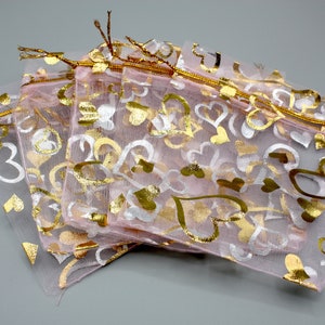 May include: A set of four pink organza gift bags with gold and white heart patterns. The bags have gold drawstring closures.