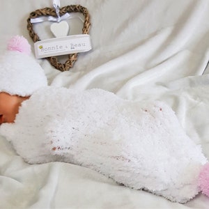 Puede incluir: Un arrullo de bebé de punto blanco con un pompón rosa en el pie y un gorro de punto blanco a juego con un pompón rosa en la parte superior. El bebé duerme plácidamente en el arrullo.