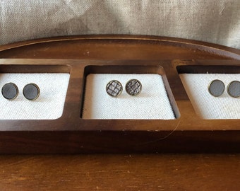 May include: Three pairs of small, round, decorative earrings displayed on a wooden tray. The earrings are made of metal and have a variety of designs, including a solid gray color, a patterned gray design, and a solid gray color.