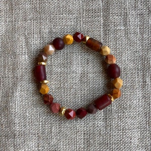 May include: A bracelet made of brown, red, and gold beads. The beads are of various shapes and sizes, and the bracelet is strung on a clear elastic cord.