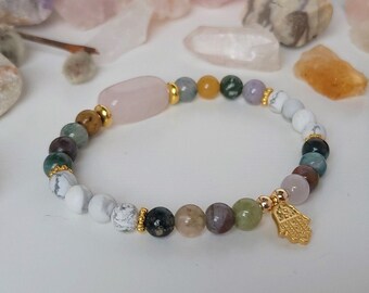 Puede incluir: Un brazalete hecho de varias piedras semipreciosas, incluyendo cuarzo rosa, jaspe y ágata. El brazalete tiene un dije de oro en forma de mano de Hamsa.