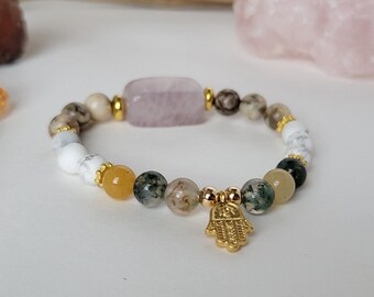 Puede incluir: Un brazalete hecho con cuentas blancas, marrones, verdes y amarillas. El brazalete tiene una piedra grande, rectangular, de color rosa claro en el centro. Un amuleto dorado en forma de mano de hamsa cuelga del brazalete.