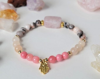 Puede incluir: Un brazalete de piedras preciosas rosa y blanco con un dije de hamsa dorado. El brazalete está hecho con cuentas redondas de varios tamaños y una piedra preciosa rosa rectangular.