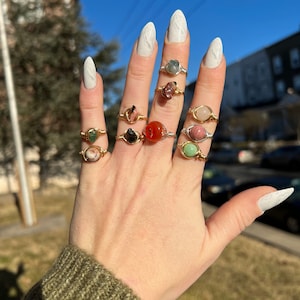 May include: A hand wearing multiple rings with different colored gemstones. The rings are made of gold wire wrapped around the stones. The stones include green, pink, red, and black.