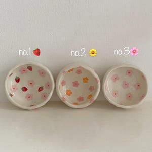 Puede incluir: Tres pequeños cuencos de cerámica blanca con diferentes patrones. El primer cuenco tiene un patrón de fresa y flor rosa. El segundo cuenco tiene un patrón de flor amarilla y flor rosa. El tercer cuenco tiene un patrón de flor rosa.