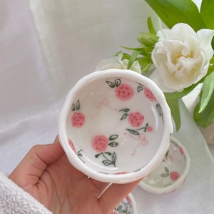 May include: A set of small, white ceramic bowls with a scalloped edge. Each bowl is decorated with pink roses and green leaves. The bowls are ideal for serving small portions of food or as decorative pieces.