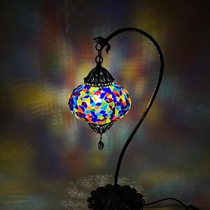 May include: A colorful mosaic table lamp with a black, curved metal stand. The lamp's shade is a vibrant mix of blue, red, yellow, and green glass pieces. The base has an ornate, floral design.