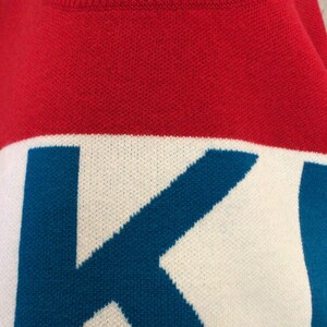 May include: Red, white, and blue knitted sweater with the word "K" in blue on the front. The sweater has a tag that says "FOKUS" and "L" for size.