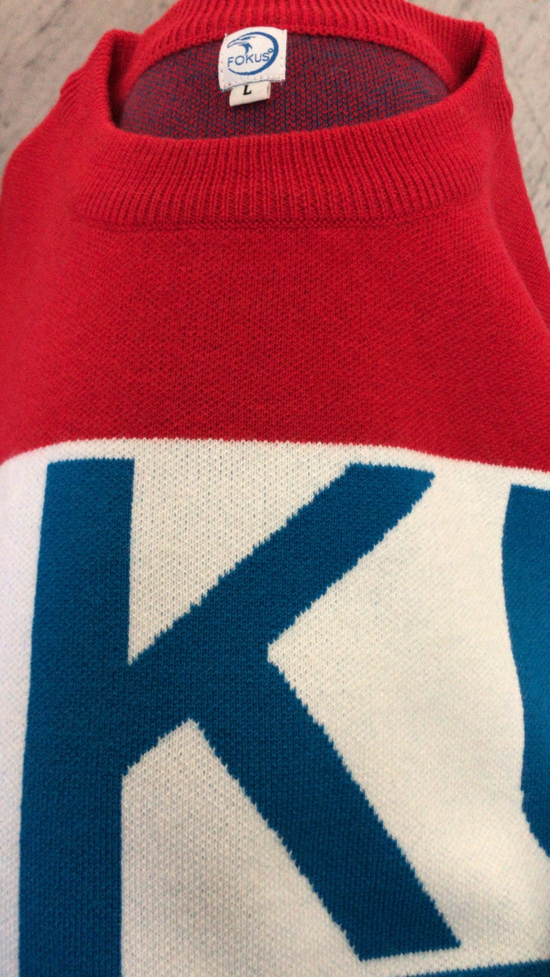 May include: Red, white, and blue knitted sweater with the word "K" in blue on the front. The sweater has a tag that says "FOKUS" and "L" for size.