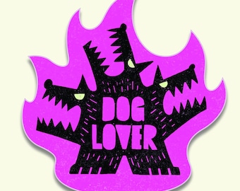May include: A sticker featuring a black illustration of a three-headed dog with the text "DOG LOVER" in white. The dog is set against a bright pink flame-shaped background, with a textured, cartoonish style.