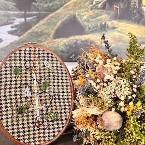 Puede incluir: Un bastidor de bordado ovalado con un marco de madera marrón y una tela con estampado de pata de gallo. El bordado presenta una espada plateada, cuentas verdes e hilo dorado. Un ramo de flores secas está a la derecha, con una pintura de paisaje al fondo.