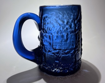 May include: A cobalt blue glass mug with a textured surface and a thick handle. The mug is designed with a raised pattern that resembles a brick wall.