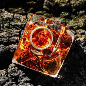 May include: A clear resin dice with a gold ring embedded inside. The dice is filled with orange and red flakes and is sitting on a dark brown textured surface.