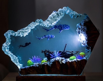 Könnte beinhalten: Eine Harzlampe mit einer durchscheinenden blauen Unterwasserszene, die zwei Walhaie, einen Taucher und ein Schiffswrack zeigt. Der Sockel besteht aus dunklem Holz und die Oberkante hat eine wellenförmige Textur. Ein internes Licht beleuchtet die Szene.